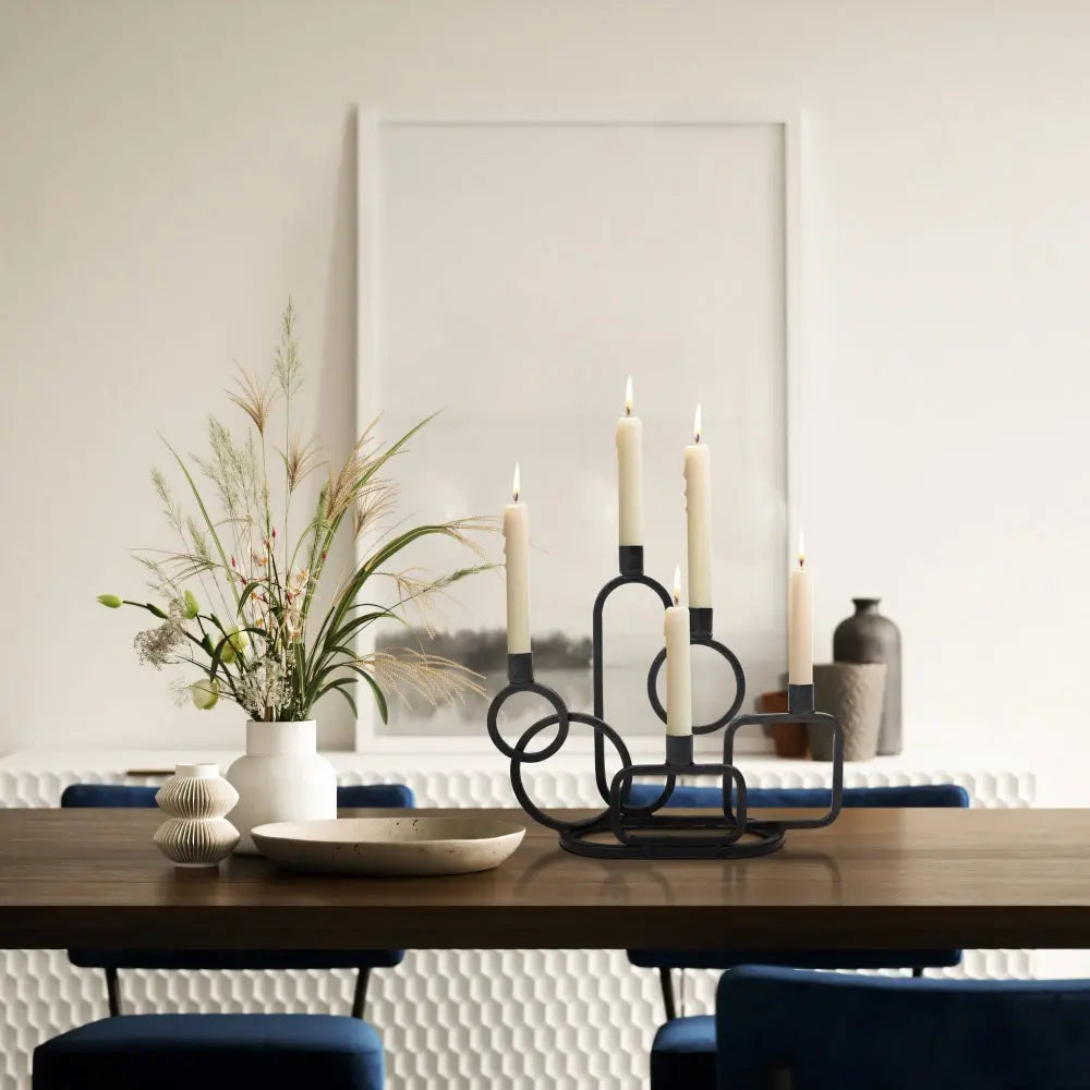 A modern dining table with a geometric black candle holder, white candles, a vase with greenery, and decorative ceramics; blurred wall art in the background.