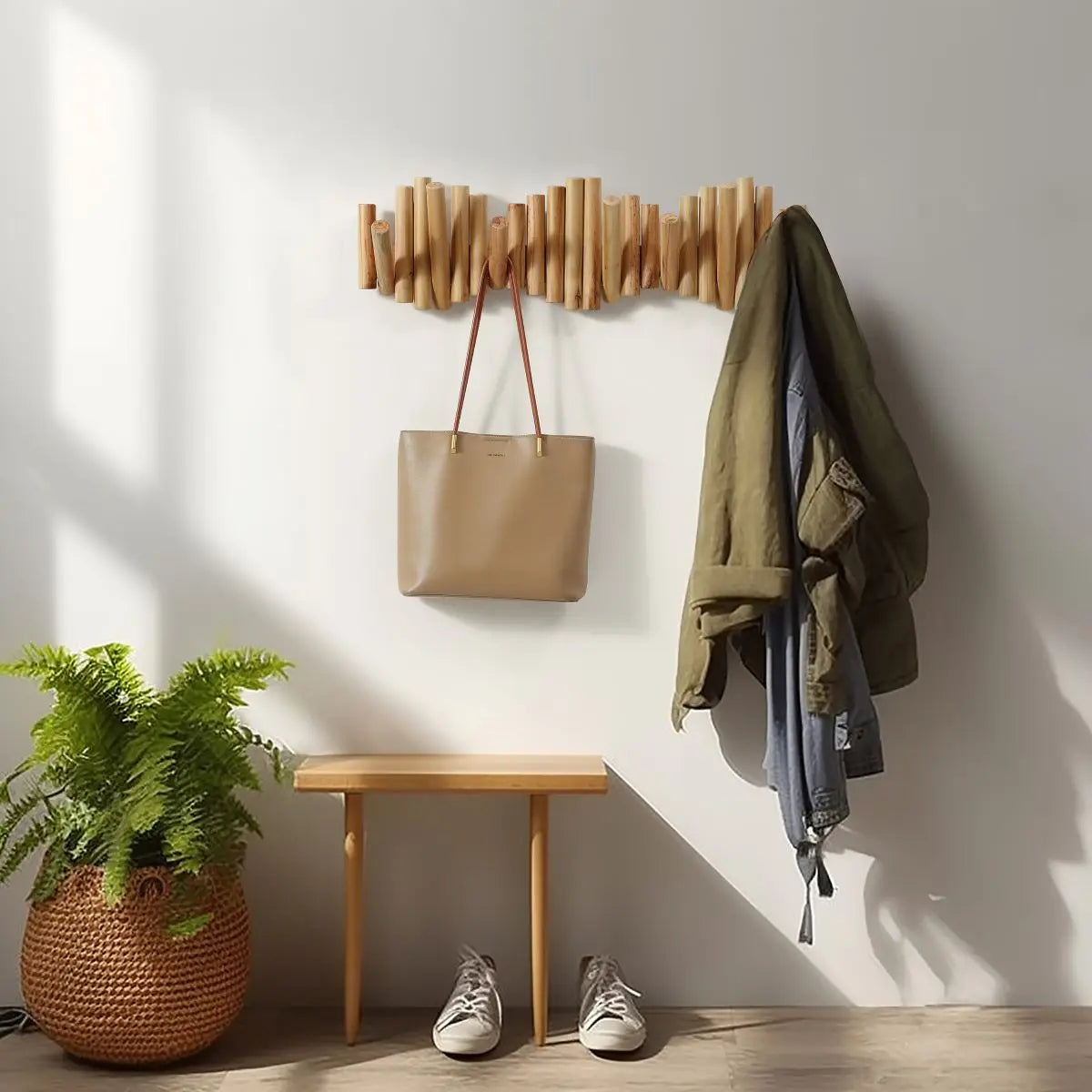 A wall-mounted wooden coat rack holds a beige tote bag and a green jacket above a small wooden bench with two pairs of shoes; a potted fern sits on the floor nearby.