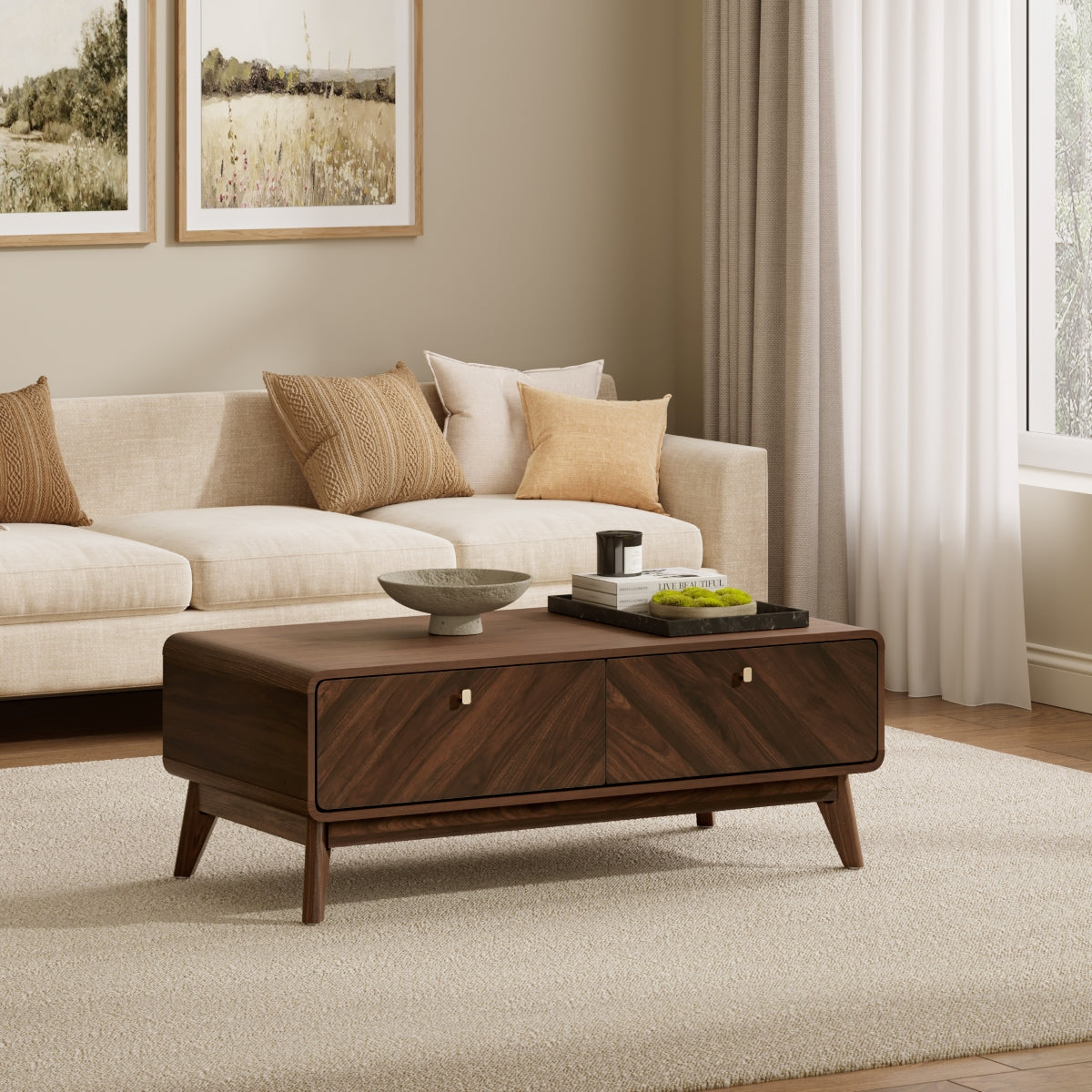 Modern living room with a beige sofa, wooden coffee table holding books and decor, and light-colored carpeting. Two landscape artworks hang on the wall above the sofa.