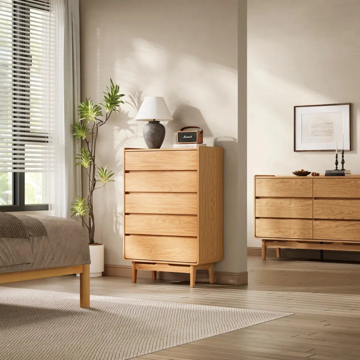 Minimalist bedroom with light wood furniture, including a dresser and bed. A potted plant, table lamp, and framed art add decor. A window with blinds allows light to enter the space.