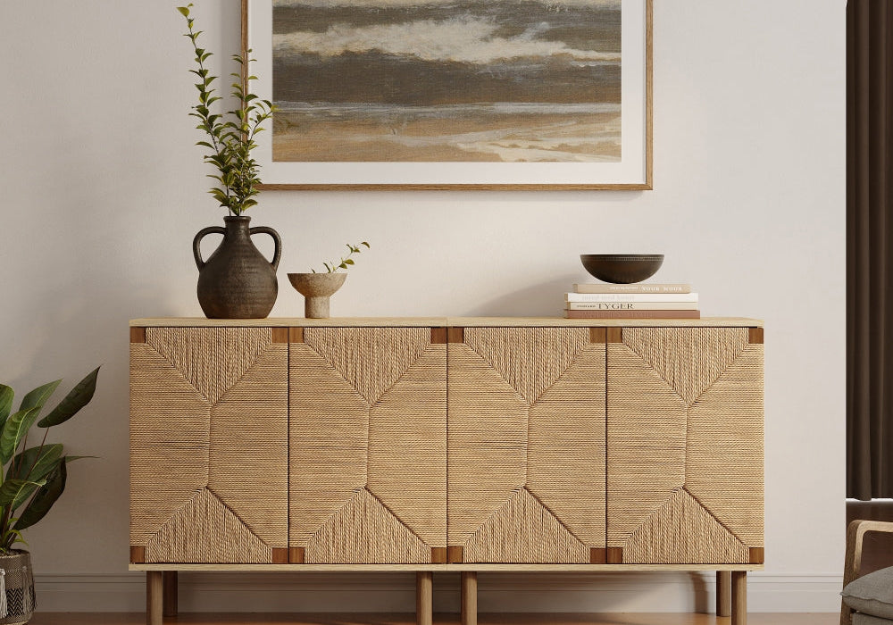 A wooden sideboard with a textured design stands against a wall. A vase with plants, a bowl on books, and a landscape painting are displayed on top. A striped rug lies on the floor.