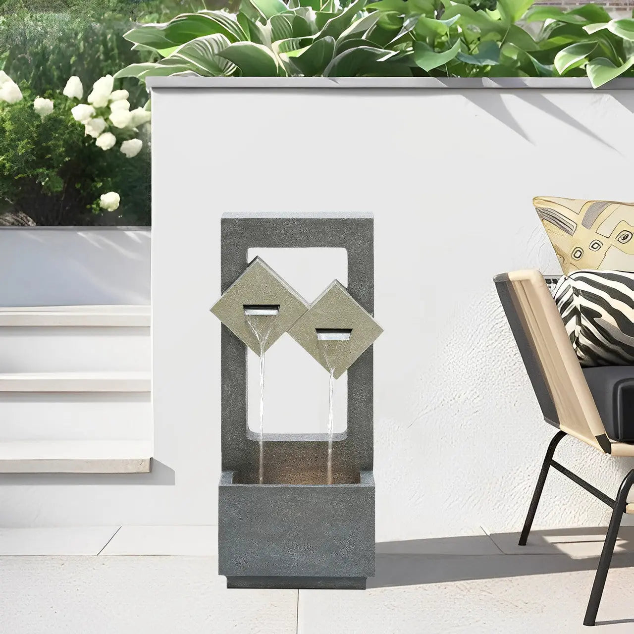 Modern outdoor fountain with two tilted square spouts, water flowing into a rectangular basin, set on a patio with plants and a cushioned chair nearby.