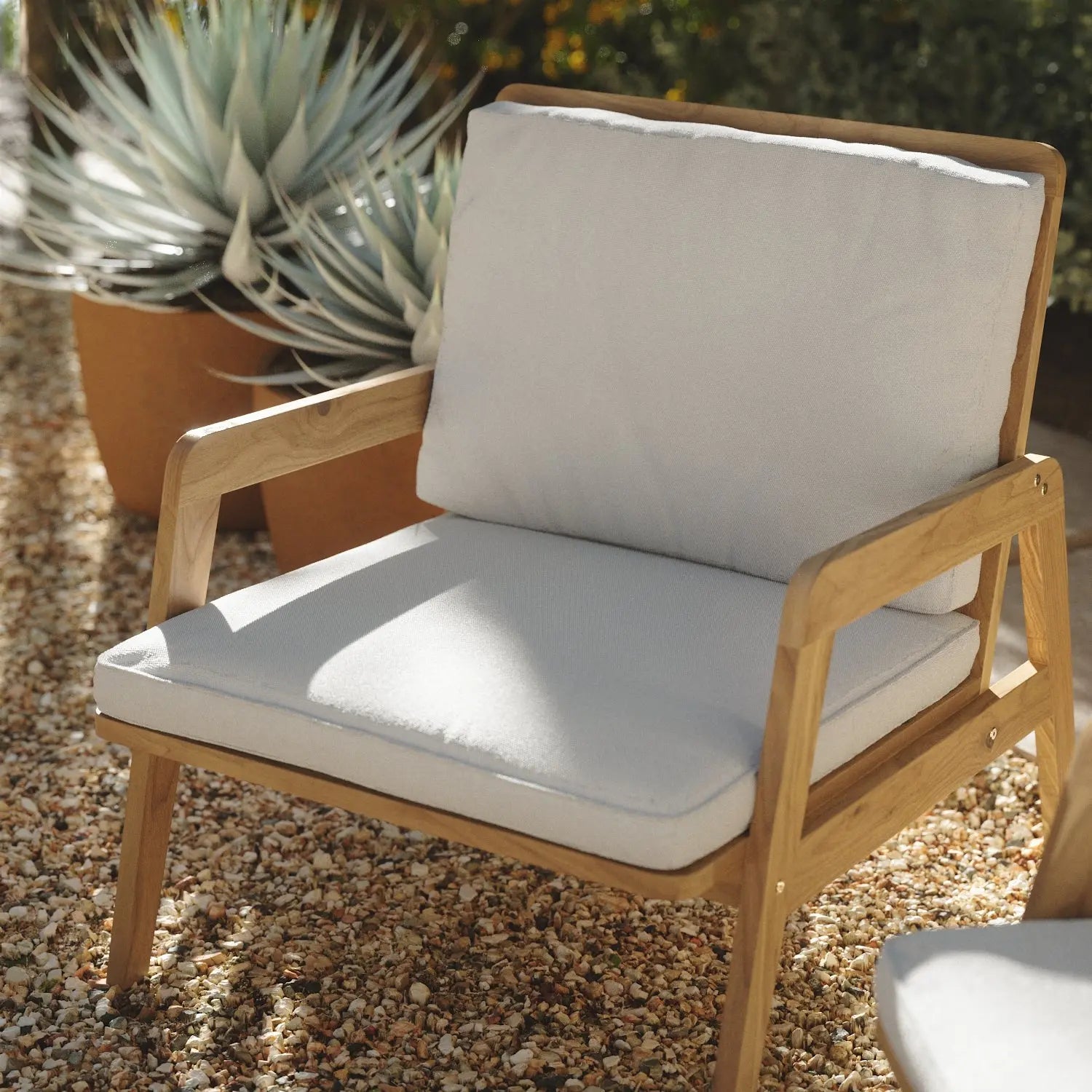 Wooden outdoor chair with white cushions on a gravel area. Large potted succulents in the background.