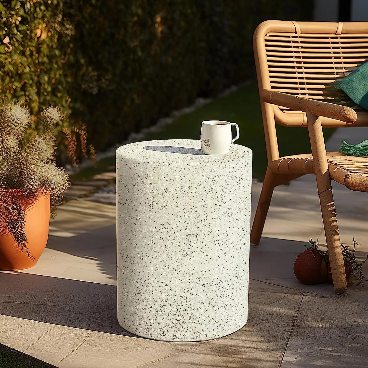 A white cylindrical side table with a coffee mug on top is placed on a patio next to a wicker chair and potted plant, with greenery in the background.