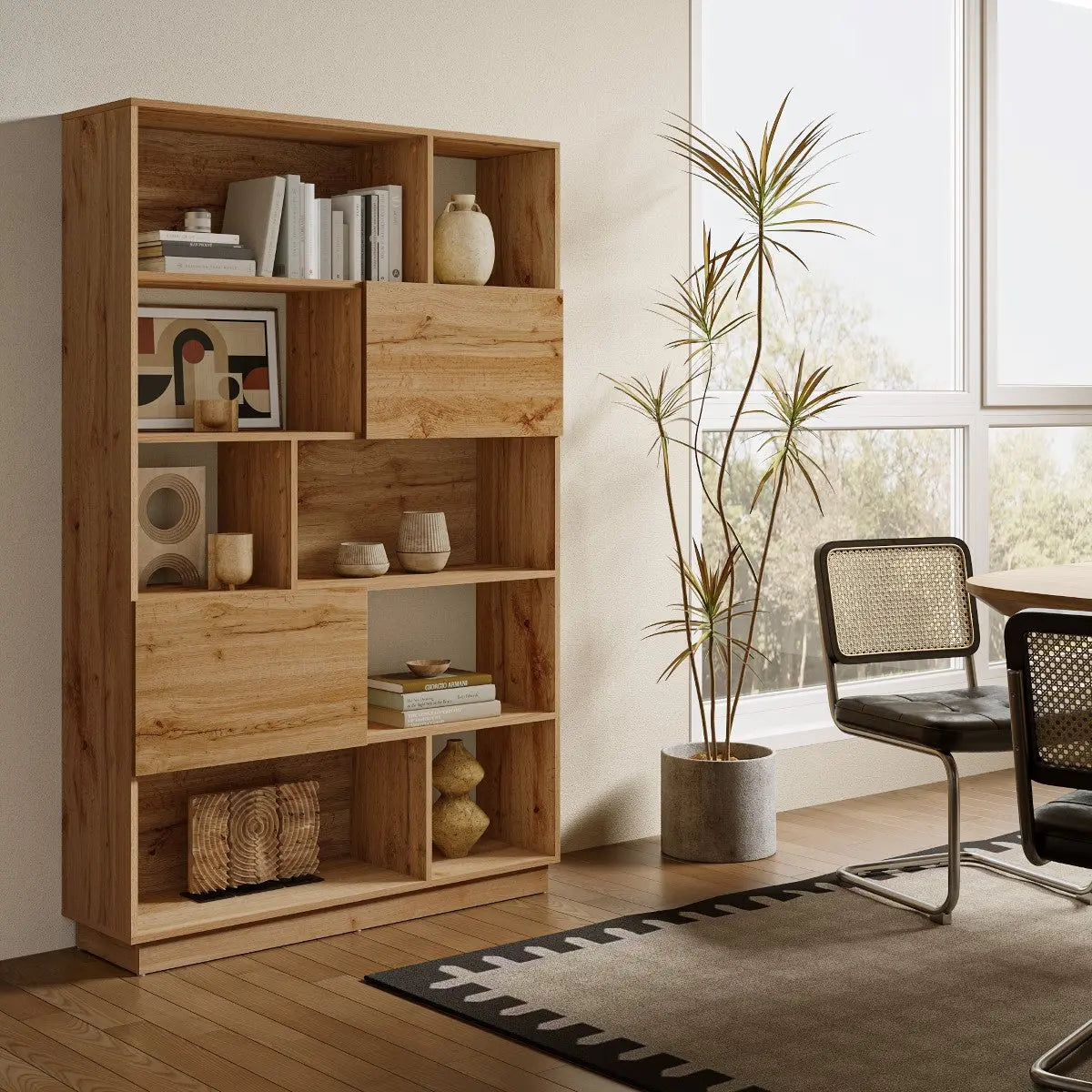 A wooden bookcase with books, vases, and decor items stands next to a potted plant, chairs, and a table in a bright, modern room with large windows.