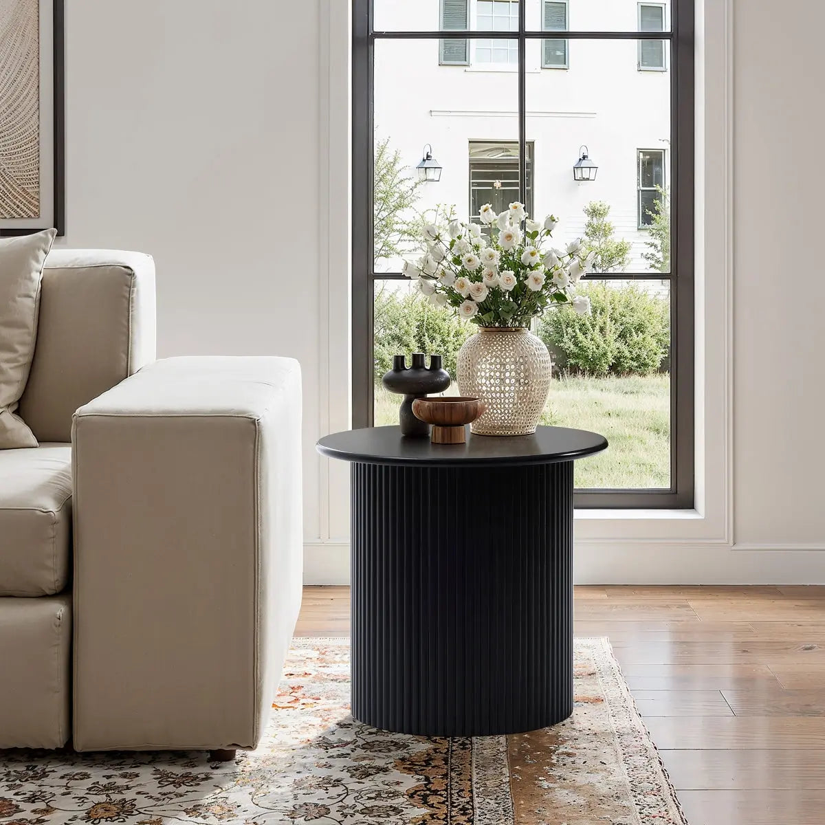 A round black side table with decorative vases and flowers stands next to a beige sofa on a patterned rug by a large window with a view of a building and greenery outside.