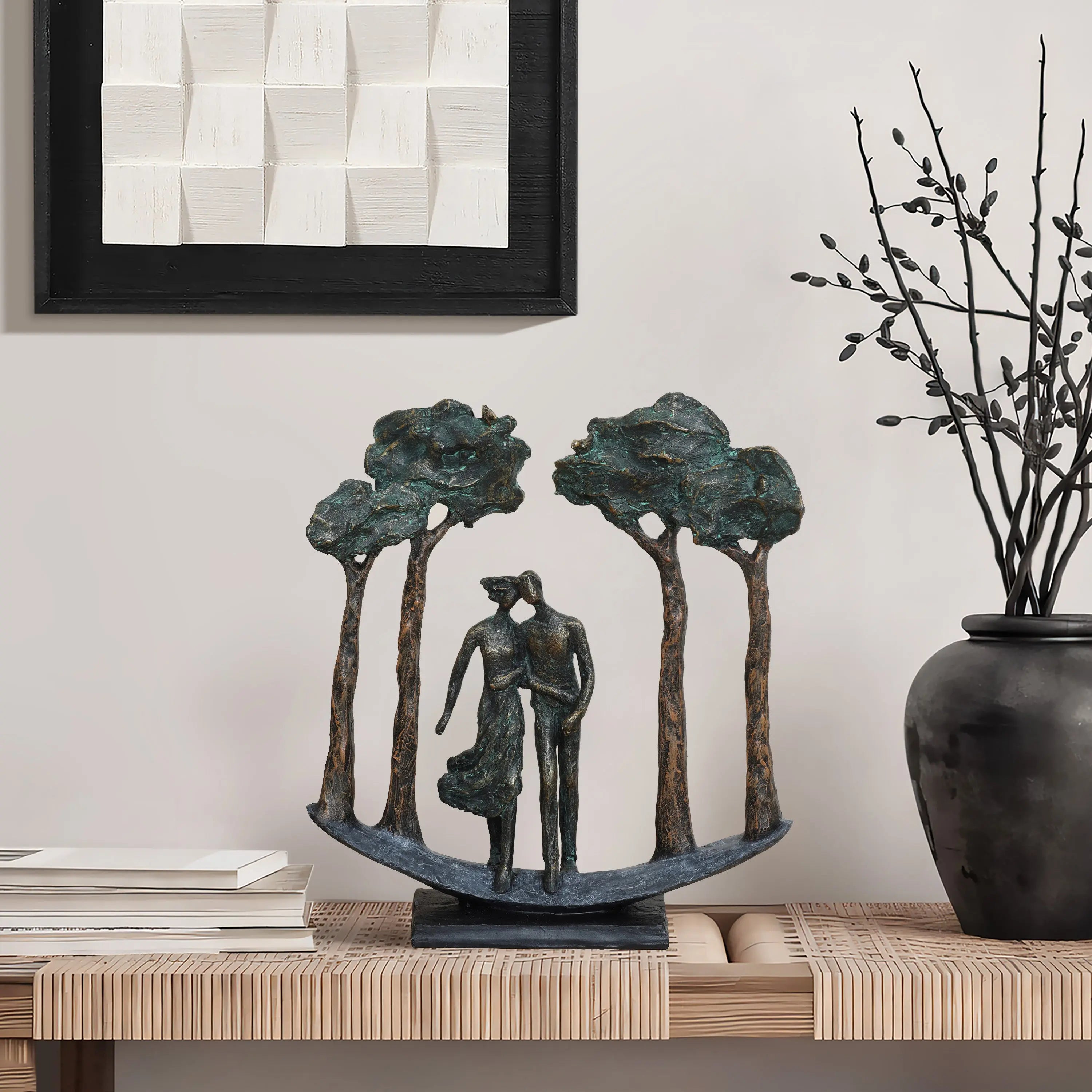 Bronze sculpture of a couple walking under trees on a table with books, next to a vase with twigs. Wall art hangs above.