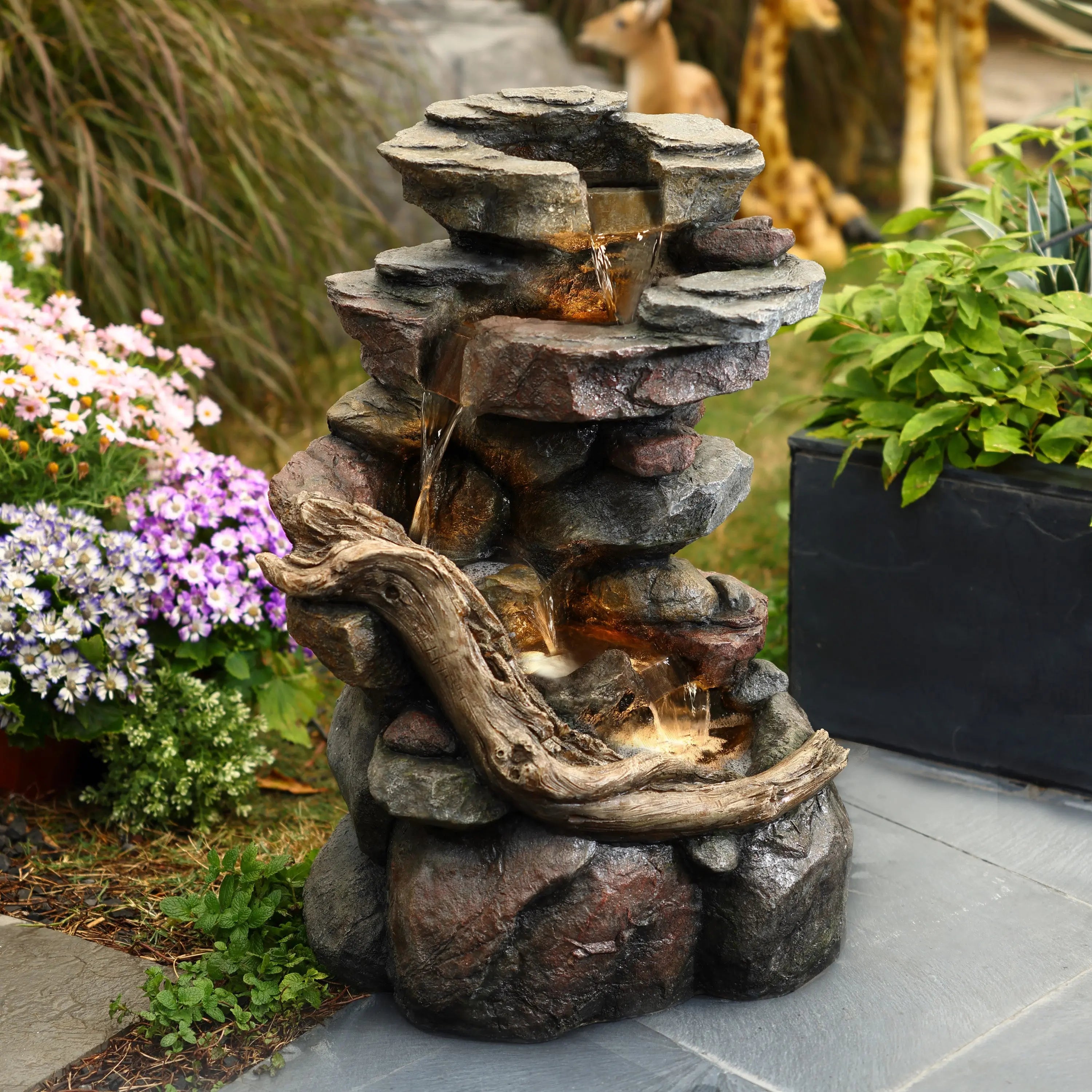 A tiered stone water fountain with cascading water and a driftwood accent sits outdoors, surrounded by flowers and green plants.