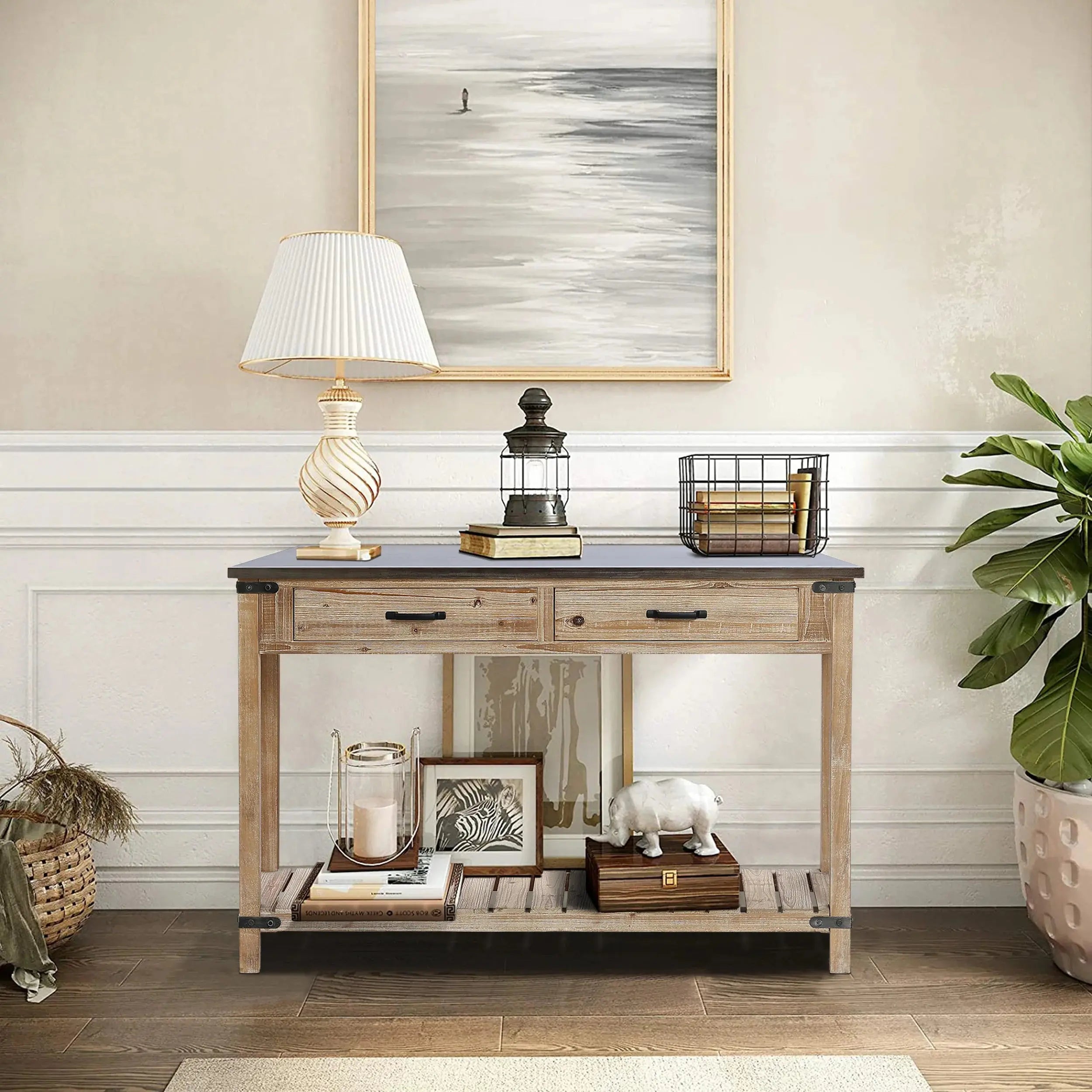 Wooden console table with a lamp, lantern, and wire basket. Lower shelf holds books and decor. Framed artwork above. Plant on the right and wicker basket on the left. White paneled wall.