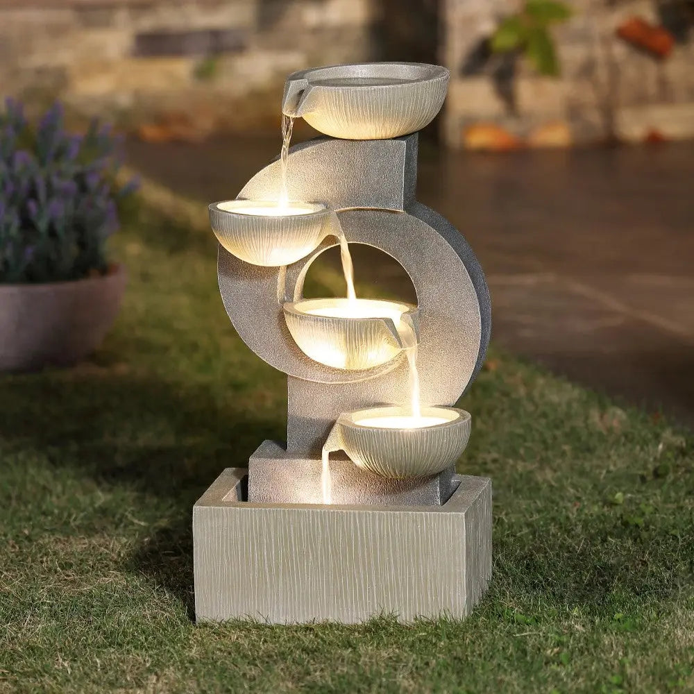 A tiered outdoor water fountain with four bowl-shaped basins, illuminated by built-in lights, stands on a grassy area near a paved walkway.