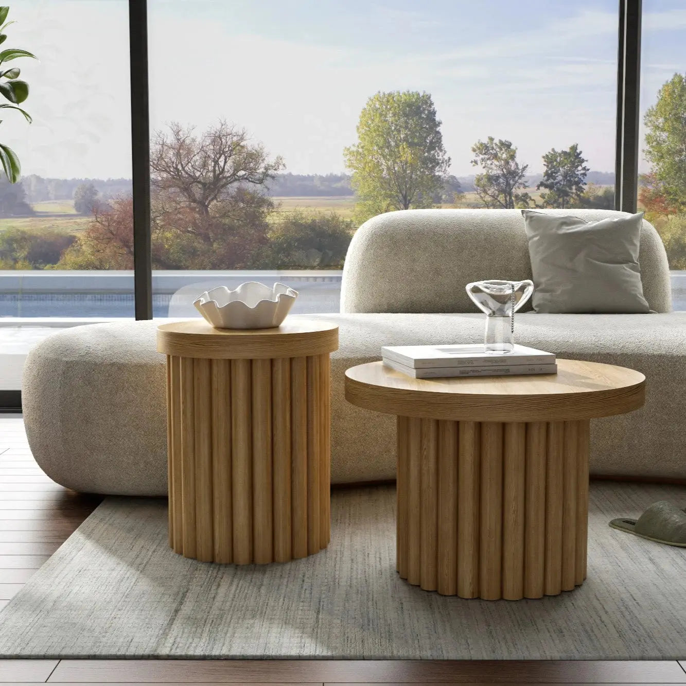 Modern living room with two round wooden side tables, a light-colored sofa, and large windows overlooking trees and a field.