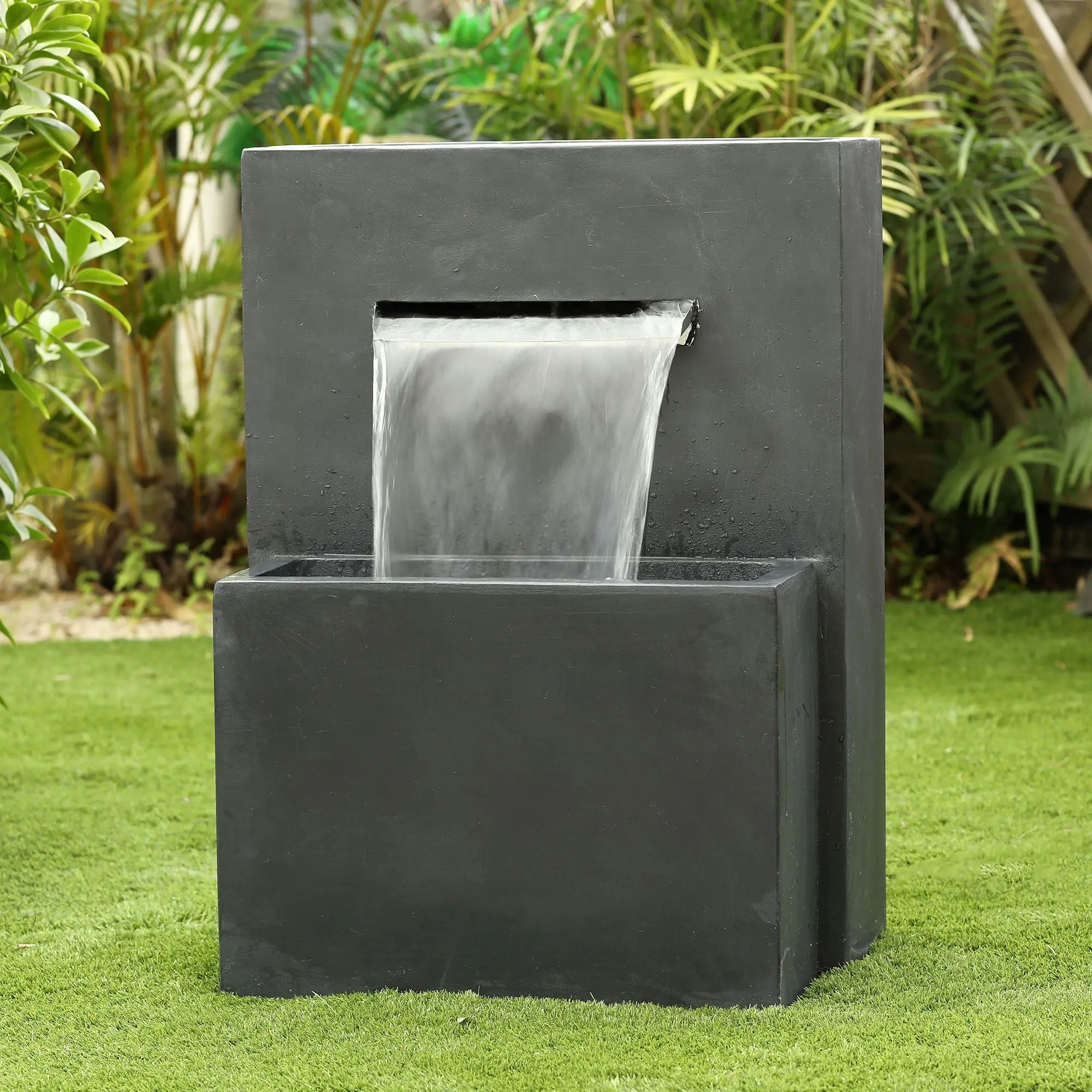 A modern rectangular black fountain with a water spout cascading into a lower basin, set on a grassy lawn with plants in the background.