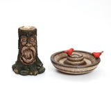 A tree trunk-shaped candle holder, resembling a brown old man, features a carved face and a decorative dish with two small red bird figurines. This charming piece serves as an outdoor decor centerpiece and adds whimsical beauty to any space.