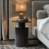 A modern bedside table with a ribbed black base and natural wood grain top elegantly holds a brown lamp with a beige shade and a decorative object. The side table complements the bed, which is partially visible under its gray bedding, enhancing the modern aesthetic.