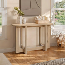 The LuxenHome Lorean natural finish contemporary console table with curved legs stands against a light wall, adorned with vases, books, and a coral ornament. Sunlight highlights the wooden floor and a woven basket beside it.