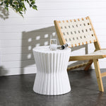 A speckled white fluted hourglass outdoor side table with a glass of water and a rolled-up newspaper sits beside a wooden chair with a woven seat, all set against a white paneled wall, accentuated by the shadow of nearby plant leaves on the floor.