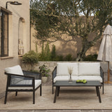 The outdoor patio features a black steel conversation set with a sofa and chair adorned in white cushions, complemented by a coffee table displaying green apples and vases. An umbrella adds shade, while a wall-mounted light illuminates the stone wall, creating an inviting atmosphere.