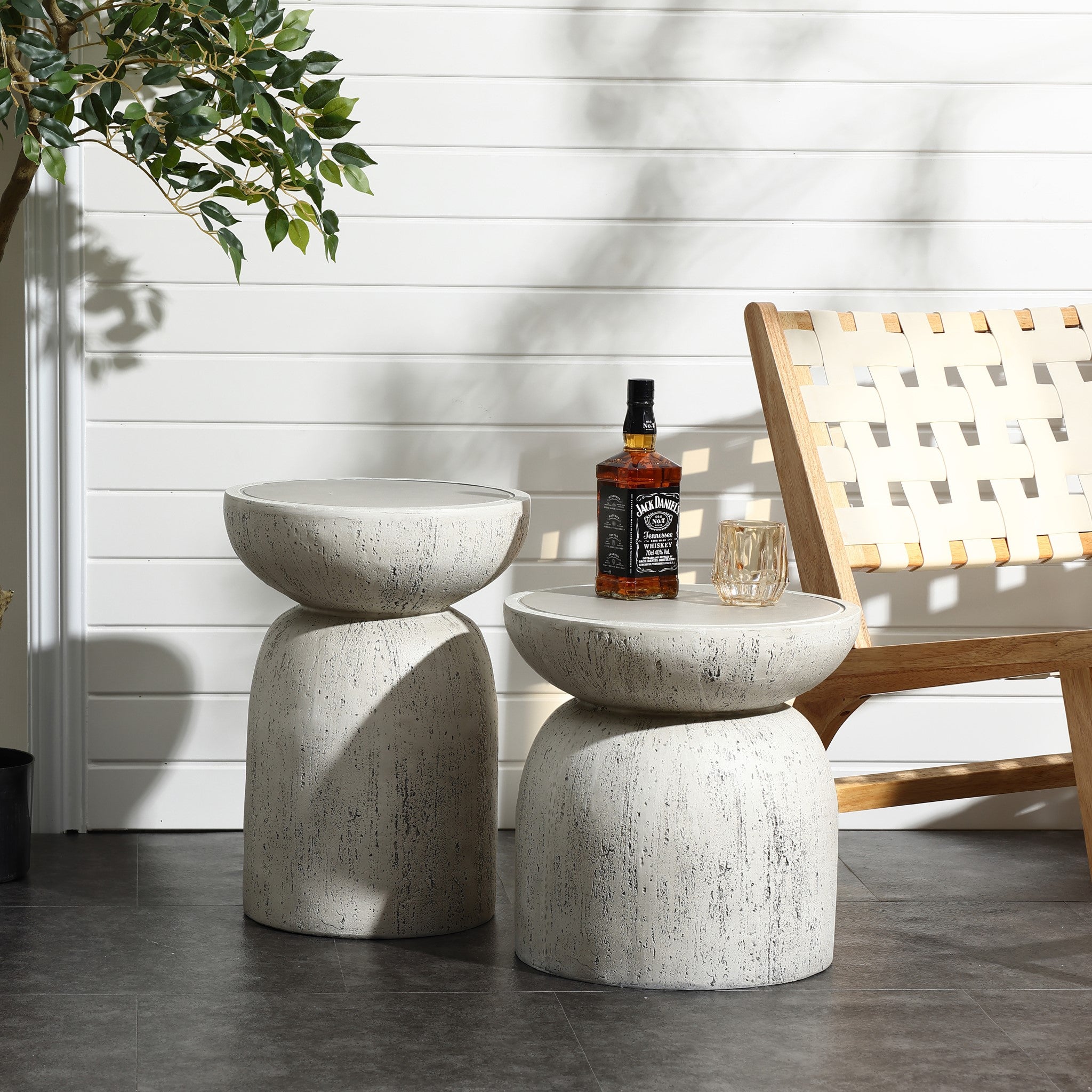 Two LuxenHome Rustic off White and gray outdoor side tables, 19.7 h, are on a patio with whiskey and a glass on one. A wooden chair with woven white seating is nearby, and a leafy plant casts shadows on a white paneled wall.