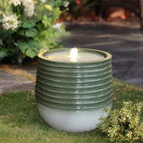 A modern fountain with a green ribbed design sits on grass, its central opening emitting water, surrounded by plants and stone flooring. LED lights illuminate the fountain, enhancing its elegant presence in the tranquil setting.