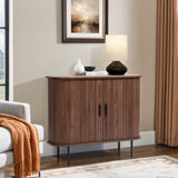 A modern living room showcases the LuxenHome Aria 2-door tambour accent cabinet in a dark oak finish, topped with a lamp, white vase, and black bowl. A framed abstract painting hangs above, an orange throw blanket drapes over the sofa, while beige curtains and a light rug complete the scene.