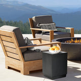 cement outdoor end table by the side of solid teak wood outdoor accent chair with grey cushions, in front of cement outdoor fire pit