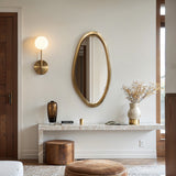 A modern living room features an elegant oval wall mirror, a wall sconce, a marble console table with decor, a stool, and a pouf.