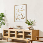 Wooden sideboard with glass doors, topped with decorative items. A potted plant and candle are on the left, with a painting of leaves featuring a gold leaf motif above. Greenery is on either side, creating a harmonious blend of nature and elegance.