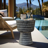 A modern poolside scene features a wicker lounge chair and the Meru outdoor side table by LuxenHome in black. Crafted from durable magnesium oxide, the table holds a potted plant, two champagne glasses, and a bottle. Clear skies meet the mountains in the background.