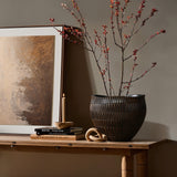 A LuxenHome dark brown textured resin round planter holds branches of small red leaves on a decorative wooden table. An abstract painting, lit candle, stacked books, and a twisted geometric wooden ornament rest nearby on a muted wall.