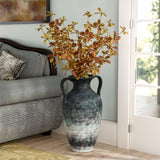 A Mottled gray ceramic tall vase by LuxenHome, 18.5 high, filled with autumn leaves, rests on a wooden floor beside a patterned couch near a large window with white curtains. The room is softly lit, showcasing minimalist elegance.