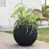 A green fern plant in a modern, durable polyethylene planter sits on a wooden deck, with wicker chairs and a white building in the background.