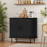 In a modern kitchen scene, the LuxenHome Aria 2-door tambour accent cabinet in black finish echoes mid-century modern style. A coffee maker, cutting board, glasses, and small potted plants rest atop it. Shelves above hold jars and plants with a potted plant nearby on the floor.