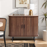 The dark oak Aria 2-door tambour accent cabinet by LuxenHome stands in a modern living room. Its finish complements the gold lamp and tray with wine glasses on top. A framed abstract art hangs above, while a chair with a woven backrest and a potted plant sit nearby.