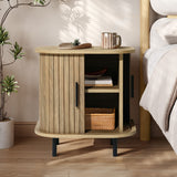 The LuxenHome Aria oak finish nightstand features a slatted sliding door and stands on four black legs, embodying mid-century modern design. With a round top supporting a small plant, wooden object, and mug, its shelves house a leaf-shaped dish and woven basket near the bed and potted plant.