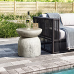 A Rustic off White and gray outdoor side table, 15.75 h, holds a bottle of white wine and glass beside a wicker outdoor sofa with a gray throw blanket. The poolside scene features a brick patio and wooden fence in the background.