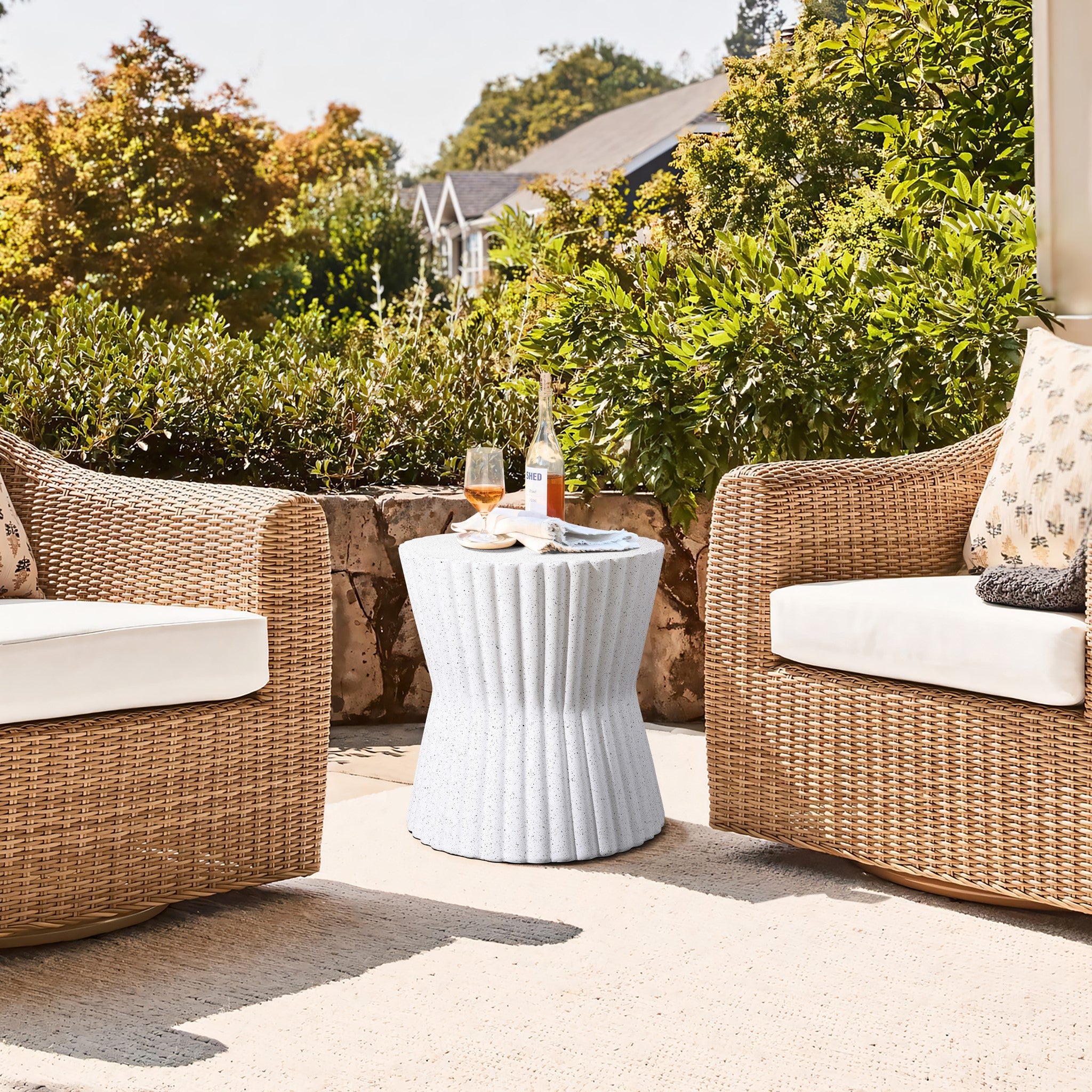 A cozy outdoor setting showcases two wicker chairs with light cushions and a speckled white fluted hourglass outdoor side table in between. On the table are a glass of orange juice and a bottle. Lush greenery and a stone wall surround the area, creating a serene atmosphere.