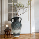 The LuxenHome Mottled gray ceramic tall vase, 18.5 h, with a weathered finish sits on a wooden floor holding sparse branches. Beside it is a small wooden stool with a modern lamp. The minimalist scene is framed by a white paneled wall and glass door behind.