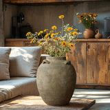 A LuxenHome rustic brown terracotta cauldron vase, 15.4 high, filled with bright yellow daisies sits on a wooden table. A beige sofa and wooden cabinet in the background add to the cozy, earthy ambiance with natural elegance.