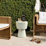 A patio scene showcases LuxenHomes Meru outdoor end table, with its beige drum-shaped design complemented by wicker chairs and white cushions. A whiskey bottle and glass adorn the minimalist table, while sandals rest on the tiles, set against a green hedge backdrop.