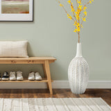 A minimalist room includes a wooden bench, white shoes, and a textured cushion. A LuxenHome Modern geometric pattern resin tall trumpet vase, 19.5 h, filled with yellow flowers stands on a patterned rug against a gray wall featuring framed art.