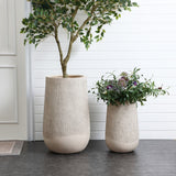 Two textured beige planters, one tall and the other shorter, stand on a dark floor against a white paneled wall. The larger round tapered planter holds a leafy tree, while the smaller displays flowers and greenery in a contemporary style.