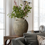 A LuxenHome rustic brown terracotta cauldron vase (15.4 h) with green leaves and pink flowers sits on a wooden table beside a gray sofa with a plaid cushion, surrounded by books and decor, while large windows illuminate the space with natural light.