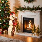 A Lighted Display snowman decoration stands by a fireplace with a fire, next to a decorated Christmas tree, gifts, and small lit trees on the hearth.