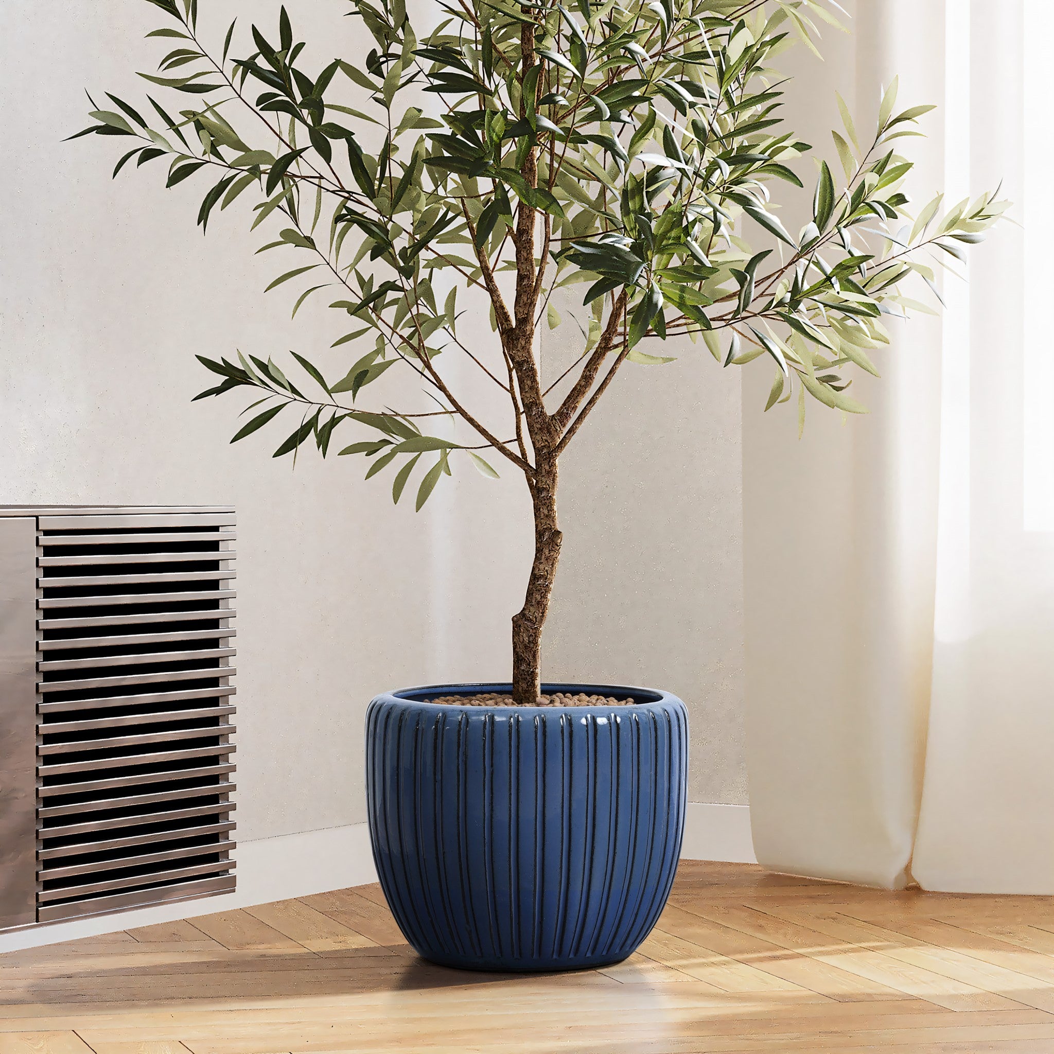 A tall green plant with thin leaves sits in a dark blue flowerpot featuring a three-dimensional vertical pattern, placed on a hardwood floor near a radiator and white curtains.