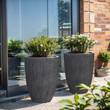 Two large, concrete-textured planters with green shrubs and white flowers stand on a patio near a glass wall and brick exterior, showcasing their UV and weather-resistant features.