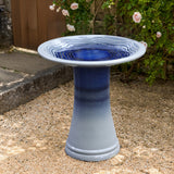 A glazed bird bath in blue and gray stands gracefully on gravel, surrounded by blooming rose bushes and a charming stone path.