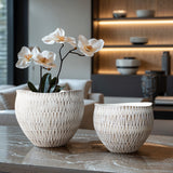 Two elegant white LuxenHome ceramic pots, featuring woven textures and geometric patterns, rest on a wooden table. The larger White textured resin round planter cradles delicate white blossoms, enhancing the modern living spaces shelves and neutral-toned decor in the background.