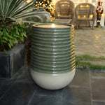 A tall candle with a ribbed design and flickering flame sits on a tiled patio, surrounded by lush plants and wicker chairs. Twinkling lights dance in the background, enhancing the serene ambiance.