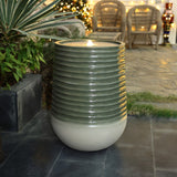 A tall candle with a ribbed design and flickering flame sits on a tiled patio, surrounded by lush plants and wicker chairs. Twinkling lights dance in the background, enhancing the serene ambiance.