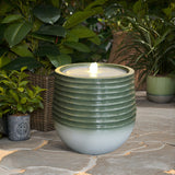 A green and white ceramic water feature with a ribbed design is set on a stone patio, surrounded by potted plants.